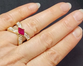 May include: A gold-toned crossover ring featuring a central marquise-cut vibrant red stone flanked by two bands encrusted with numerous small clear white stones. The design forms an X-shape across the finger, showcasing a polished metal finish.