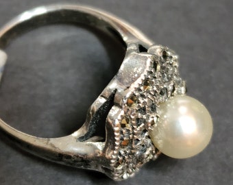 May include: A silver ring featuring a central round white pearl set within a textured, darkened metal frame. The band is smooth and polished, while the setting surrounding the pearl displays a detailed, granular surface with small dark accents.