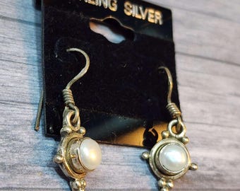 May include: A pair of sterling silver earrings with a pearl center. The earrings are on a black display card with the words "STERLING SILVER" in gold lettering. The earrings have a hook closure and a decorative design around the pearl.