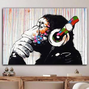 Könnte beinhalten: Großer Leinwanddruck mit einem schwarz-weißen Schimpansen, der bunte Kopfhörer trägt. Der Schimpanse hat eine Hand vor dem Mund und farbige Farbspritzer im Gesicht. Der Hintergrund hat vertikale Farbstreifen.