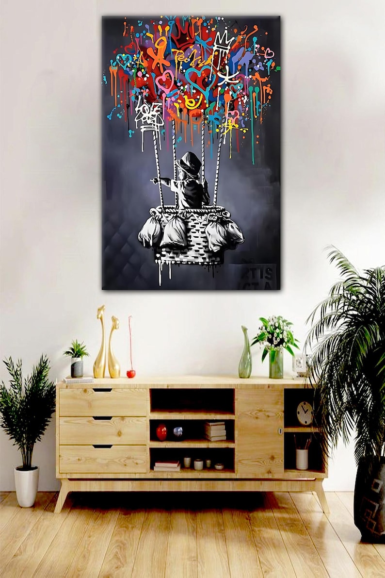 May include: A black and white painting of a child in a basket, holding onto colourful balloon-like shapes. The child is wearing a hooded jacket and is pointing to the right. The background is a dark grey with drips of paint.