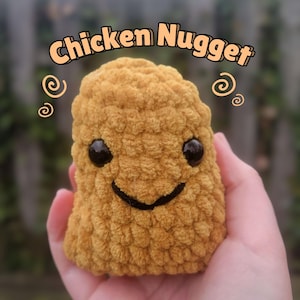 May include: A crocheted chicken nugget plush toy with a smiling face. The toy is yellow and brown and has a textured surface.