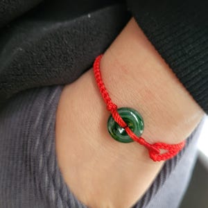 Puede incluir: Una pulsera de hilo rojo con un colgante de donut de jade verde oscuro. La pulsera se lleva en la muñeca, con una manga negra y ropa gris en el fondo. El jade es de un color verde intenso y rico.