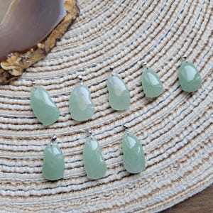 Può includere: Sette pendenti di giada verde chiaro con bails color argento sono disposti su una superficie beige intrecciata. I pendenti hanno forme organiche irregolari e variano leggermente di dimensioni. Una sezione di una pietra lucida, marrone e beige è visibile in alto a sinistra.