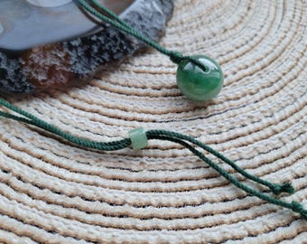 Può includere: Una collana con pendente di giada con una perla di giada verde, rotonda e lucida. La perla è sospesa a un cordoncino verde scuro con un nodo scorrevole regolabile. Lo sfondo è una superficie beige intrecciata.