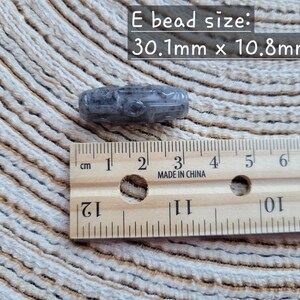 Puede incluir: Una cuenta gris tallada, de 3,01 cm x 1,08 cm, junto a una regla de madera marcada en cent&iacute;metros. El texto "E bead size: 30.1mm x 10.8mm" se muestra encima de la cuenta. La regla est&aacute; marcada "MADE IN CHINA".