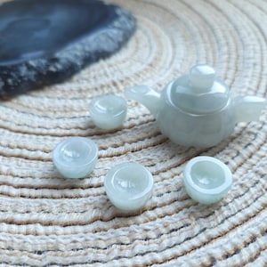 Könnte beinhalten: Ein Miniatur-Teeservice aus Jade in blassgrüner Farbe, bestehend aus einer Teekanne und vier Tassen. Das Set wird auf einer strukturierten, hellfarbigen Oberfläche präsentiert, mit einem dunklen, unregelmäßig geformten Stein im Hintergrund. Die Teekanne hat einen runden Körper und einen kleinen Griff.