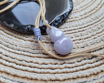 Puede incluir: Un colgante de calabaza de jade lavanda sobre un cordón beige. El amuleto en forma de calabaza tiene una superficie lisa y pulida y está suspendido de un cordón trenzado con un acento azul claro. El fondo presenta una estera tejida y una superficie oscura y moteada.
