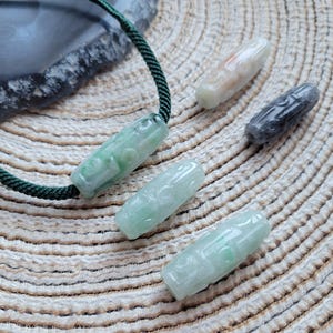 Puede incluir: Varios colgantes de jade en tonos verdes, blancos y grises. Uno est&aacute; ensartado en un cord&oacute;n verde. Los colgantes cil&iacute;ndricos tienen dise&ntilde;os tallados. El fondo es una superficie clara y texturizada. Medidas en cent&iacute;metros.