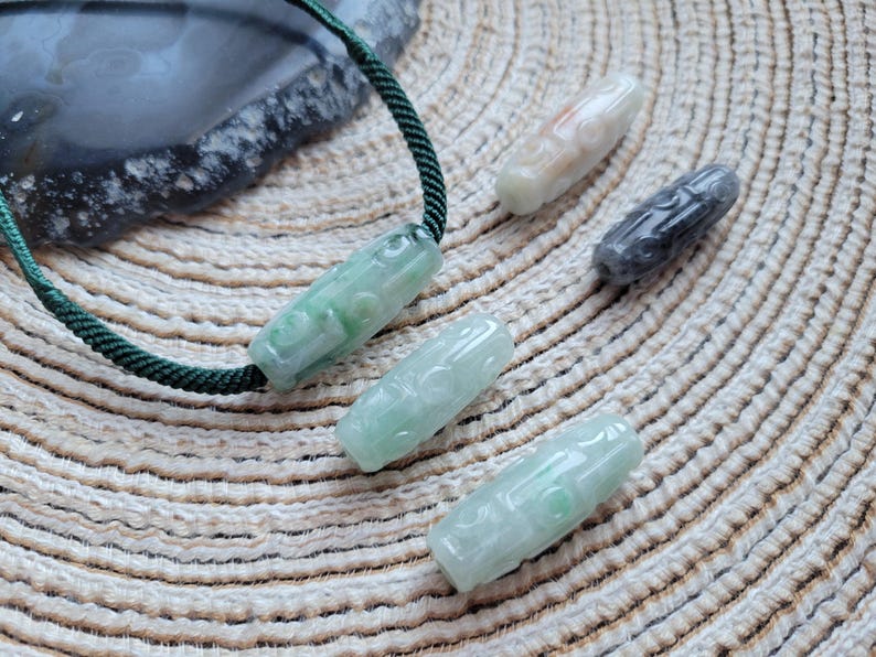 Puede incluir: Varios colgantes de jade en tonos verdes, blancos y grises. Uno est&aacute; ensartado en un cord&oacute;n verde. Los colgantes cil&iacute;ndricos tienen dise&ntilde;os tallados. El fondo es una superficie clara y texturizada. Medidas en cent&iacute;metros.