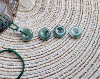 Può includere: Quattro ciondoli di giada con un cordino verde scuro, esposti su una superficie beige strutturata. I ciondoli sono circolari, con un foro centrale e diverse sfumature di verde. Un ciondolo è attaccato a un cordino verde.