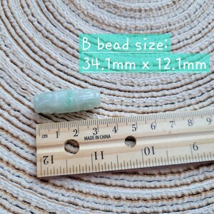 Puede incluir: Una cuenta de jade verde claro con detalles tallados, que mide aproximadamente 3,4 cm de largo y 1,2 cm de ancho. Una regla de madera con marcas de cent&iacute;metros est&aacute; al lado de la cuenta. El texto "B bead size: 34.1mm x 12.1mm" est&aacute; en la imagen.