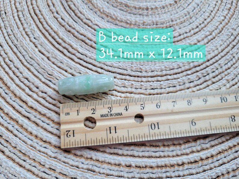 Puede incluir: Una cuenta de jade verde claro con detalles tallados, que mide aproximadamente 3,4 cm de largo y 1,2 cm de ancho. Una regla de madera con marcas de cent&iacute;metros est&aacute; al lado de la cuenta. El texto "B bead size: 34.1mm x 12.1mm" est&aacute; en la imagen.