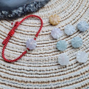 May include: A red braided bracelet with a light purple, hexagonal charm and gold accents. Several other hexagonal charms in various pastel colors are scattered nearby. The charms have a detailed, circular design. The background is a textured, beige surface.