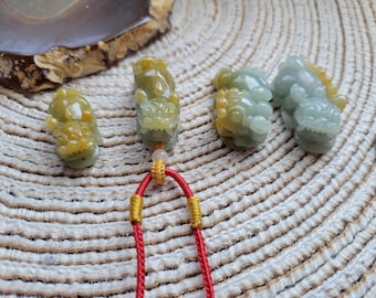Puede incluir: Cuatro colgantes de jade con forma de dragón en tonos verdes y amarillos. Los colgantes están tallados con detalles intrincados y están sujetos a un cordón rojo con detalles dorados. Se muestran sobre una superficie texturizada blanquecina.