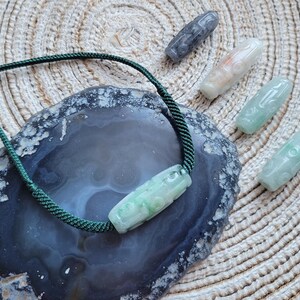 Puede incluir: Un collar de jade con un cord&oacute;n verde y un colgante de jade tallado. Varias otras piezas de jade talladas en tonos verdes, grises y blancos est&aacute;n dispersas alrededor del collar sobre una superficie texturizada. Las piezas de jade miden aproximadamente 5-7 cm de largo.