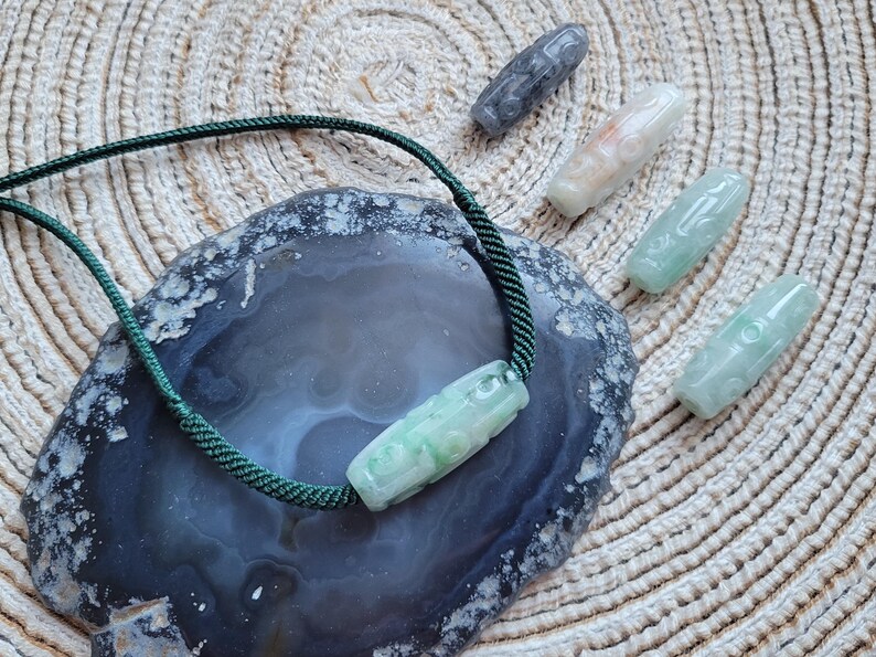 Puede incluir: Un collar de jade con un cord&oacute;n verde y un colgante de jade tallado. Varias otras piezas de jade talladas en tonos verdes, grises y blancos est&aacute;n dispersas alrededor del collar sobre una superficie texturizada. Las piezas de jade miden aproximadamente 5-7 cm de largo.