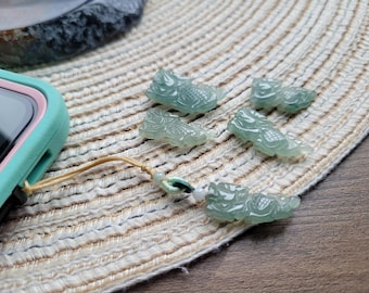 May include: A collection of translucent, light green jade dragon charms. The charms are intricately carved and arranged on a textured, cream-colored surface. A smartphone with a teal case and a lanyard is visible on the left.