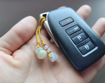 May include: A black car key fob with silver accents and a decorative gold-colored cord with jade-like charms. The key fob has buttons labeled "HOLD", a lock symbol, and a car symbol.