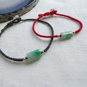 May include: Two adjustable cord bracelets, one in black and one in red, each featuring a cylindrical green jade bead centered between two small gold-toned metal beads. The bracelets have a simple knotted closure design.