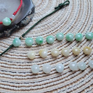 May include: A collection of jade beads in various shades of green, yellow, and white. Some beads are strung on red and green cords. The beads are displayed on a textured, light brown surface, showcasing their natural colors and shapes.
