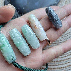 Puede incluir: Varios colgantes de jade tallado en varios colores, incluyendo verde, blanco y gris. Los colgantes cil&iacute;ndricos presentan dise&ntilde;os intrincados y se muestran en una mano. Una cuerda verde est&aacute; unida a algunos de ellos.