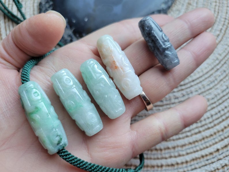 Puede incluir: Varios colgantes de jade tallado en varios colores, incluyendo verde, blanco y gris. Los colgantes cil&iacute;ndricos presentan dise&ntilde;os intrincados y se muestran en una mano. Una cuerda verde est&aacute; unida a algunos de ellos.