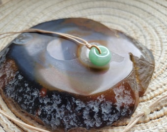 Puede incluir: Un colgante de donut de jade verde claro sobre un cordón beige. El colgante es redondo con un agujero en el centro. Descansa sobre una piedra pulida multicolor con tonos marrones, grises y blancos.