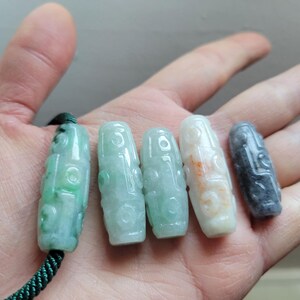 Puede incluir: Se muestran cinco colgantes de jade en varios tonos de verde, blanco y gris. Los colgantes tienen un dise&ntilde;o tallado y se sostienen en una mano. Un colgante est&aacute; sujeto a un cord&oacute;n verde.