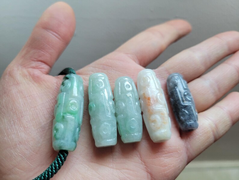 Puede incluir: Se muestran cinco colgantes de jade en varios tonos de verde, blanco y gris. Los colgantes tienen un dise&ntilde;o tallado y se sostienen en una mano. Un colgante est&aacute; sujeto a un cord&oacute;n verde.
