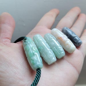 Puede incluir: Cinco colgantes de jade en varios tonos de verde, blanco y gris se muestran en una mano. Cada colgante es cil&iacute;ndrico con detalles tallados y ensartado en un cord&oacute;n verde oscuro. Los colgantes miden aproximadamente 3,8 cm de largo.