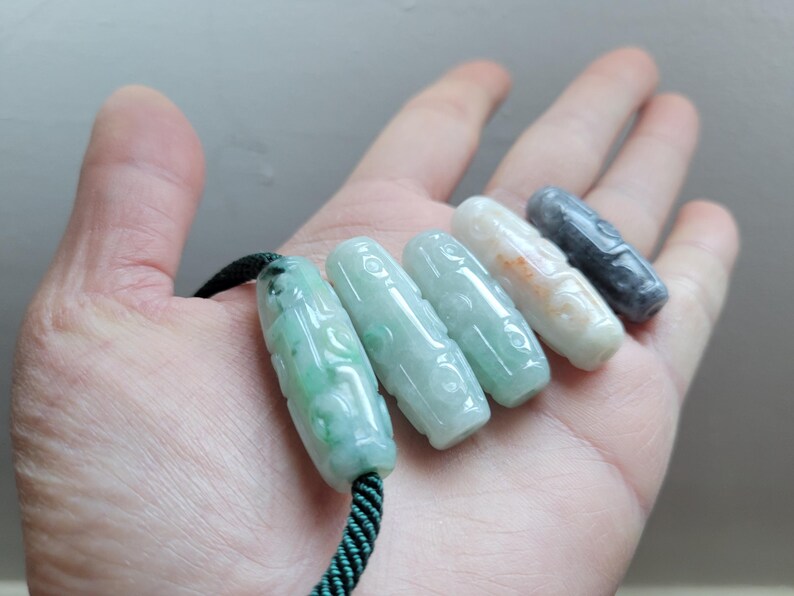 Puede incluir: Cinco colgantes de jade en varios tonos de verde, blanco y gris se muestran en una mano. Cada colgante es cil&iacute;ndrico con detalles tallados y ensartado en un cord&oacute;n verde oscuro. Los colgantes miden aproximadamente 3,8 cm de largo.
