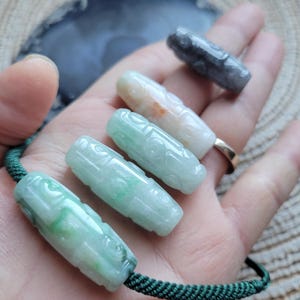 Puede incluir: Una colecci&oacute;n de colgantes de jade tallados en varios colores, incluyendo verde, blanco y gris. Los colgantes son cil&iacute;ndricos con dise&ntilde;os intrincados y est&aacute;n ensartados en un cord&oacute;n verde. Los colgantes se muestran en una mano.