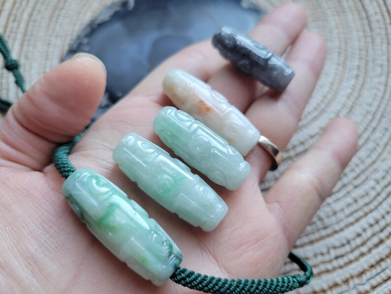 Puede incluir: Una colecci&oacute;n de colgantes de jade tallados en varios colores, incluyendo verde, blanco y gris. Los colgantes son cil&iacute;ndricos con dise&ntilde;os intrincados y est&aacute;n ensartados en un cord&oacute;n verde. Los colgantes se muestran en una mano.