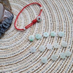 May include: A red string bracelet with a flower-shaped jade charm and several loose jade flower beads. The beads are light green and white, scattered on a textured, beige surface.
