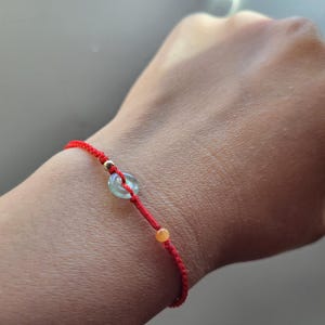 May include: Red string bracelet with a gold bead and a light blue, circular bead. The bracelet also features a small, orange bead. The bracelet is worn on a wrist.