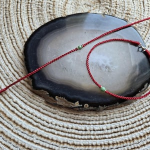 May include: Two red cord bracelets with green beads and gold-colored clasps are displayed on a dark gray and white stone. One bracelet is a simple cord, and the other is a circular bracelet.