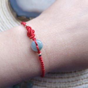 Puede incluir: Pulsera de hilo rojo con una cuenta azul verdoso claro y detalles dorados. La pulsera tiene un diseño de nudo decorativo. Una joya sencilla y elegante.