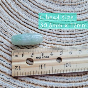 Puede incluir: Una cuenta de jade verde claro, de forma ovalada, con detalles tallados. La cuenta mide aproximadamente 3,1 cm de largo por 1,2 cm de ancho. El texto en la imagen dice "C bead size 30.6mm x 12mm". Una regla de madera est&aacute; colocada junto a la cuenta.