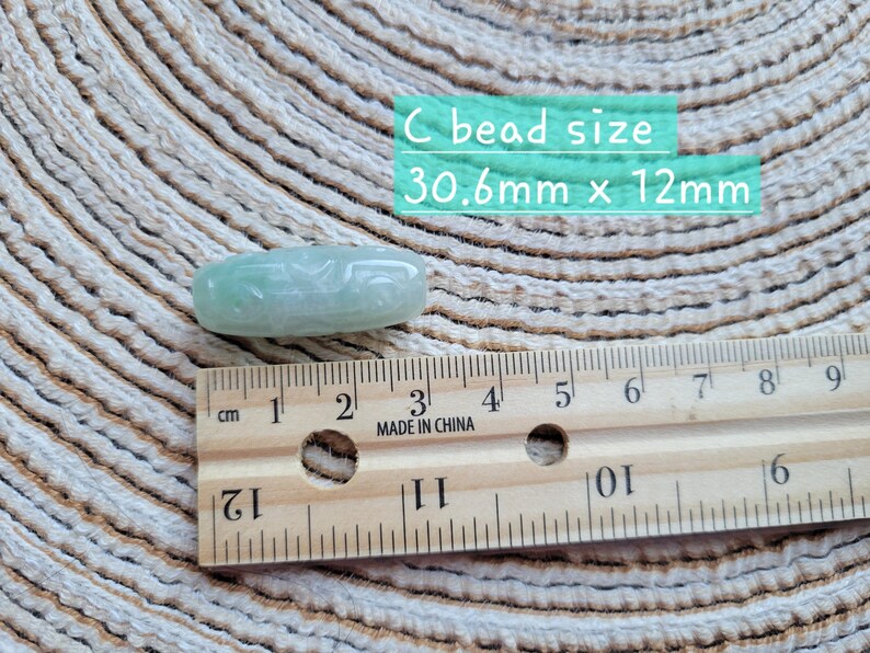 Puede incluir: Una cuenta de jade verde claro, de forma ovalada, con detalles tallados. La cuenta mide aproximadamente 3,1 cm de largo por 1,2 cm de ancho. El texto en la imagen dice "C bead size 30.6mm x 12mm". Una regla de madera est&aacute; colocada junto a la cuenta.