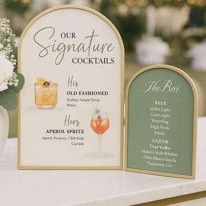 May include: Two arched signs with gold frames. One sign reads "Our Signature Cocktails" and lists "His" Old Fashioned and "Hers" Aperol Spritz. The other sign says "The Bar" and lists beer and liquor options.