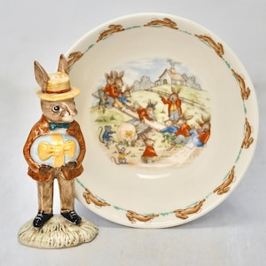 May include: A ceramic figurine of a rabbit in a brown coat, hat, and bow tie, holding a blue and yellow gift. Beside it is a cream-colored bowl with a rabbit scene and a border of brown rabbits.