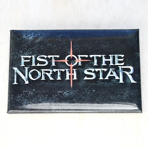 May include: A rectangular magnet featuring the text "FIST OF THE NORTH STAR" in white, stylized lettering. The background is a dark, textured blue and black. A red compass-like design is centered above the text.