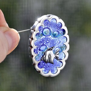 May include: A silver oval pendant with a blue and white glass design featuring a fish and bubbles. The pendant has a scalloped edge.