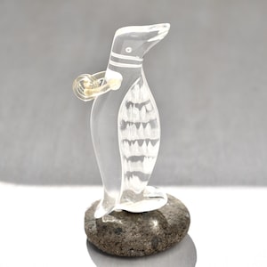 May include: A clear glass penguin figurine with white painted details, standing on a small gray rock. The penguin has a ring around its neck.