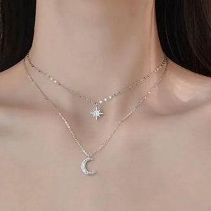 May include: A silver-coloured, layered necklace with a star pendant on the upper layer and a crescent moon pendant on the lower layer. Both pendants are adorned with small, sparkling details. The necklace is shown against a plain backdrop.