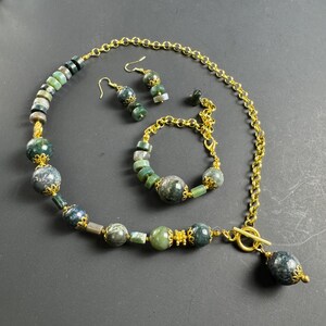 Può includere: Un set di gioielli in agata verde e oro con una collana, un braccialetto e orecchini. La collana ha una chiusura a scorrimento e un grande ciondolo in agata. Il braccialetto e gli orecchini sono anche realizzati con perline di agata e dettagli dorati.