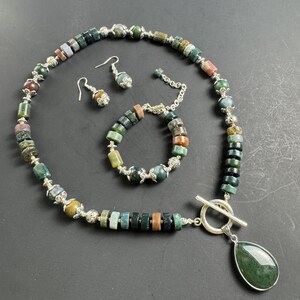 Op de afbeelding: Een groene, bruine en grijze agaatstenen set met ketting, armband en oorbellen met zilveren accenten. De ketting heeft een traanvormige hanger.