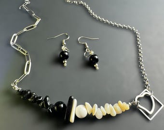 May include: A silver necklace and earring set with black and white stone accents. The necklace features a chain and a pendant with a heart-shaped design. The earrings have black beads. The jewelry is set against a dark gray background.