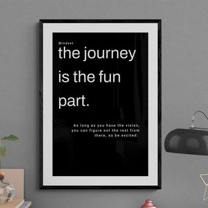 May include: A framed black and white print with the text "the journey is the fun part." The print is on a desk with a lamp, small red jars, and a plant in a glass vase. The lamp has a gold star-shaped decoration.